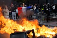 Foto: Seorang demonstran membentangkan bendera Prancis di dekat kobaran api yang membakar barikade jalanan saat aksi unjuk rasa besar-besaran menuntut penurunan biaya hidup berujung ricuh di Paris.
