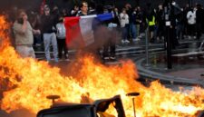 Foto: Seorang demonstran membentangkan bendera Prancis di dekat kobaran api yang membakar barikade jalanan saat aksi unjuk rasa besar-besaran menuntut penurunan biaya hidup berujung ricuh di Paris.